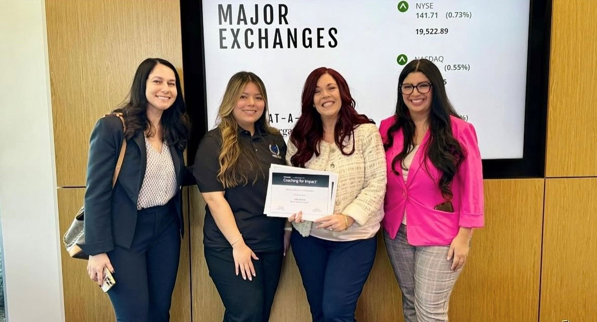 A group of women is holding a certificate, showcasing their achievement. They are indoors, smiling and dressed casually in clothing like jeans. The setting appears to have a wall in the background, and they stand together as a team celebrating their success.