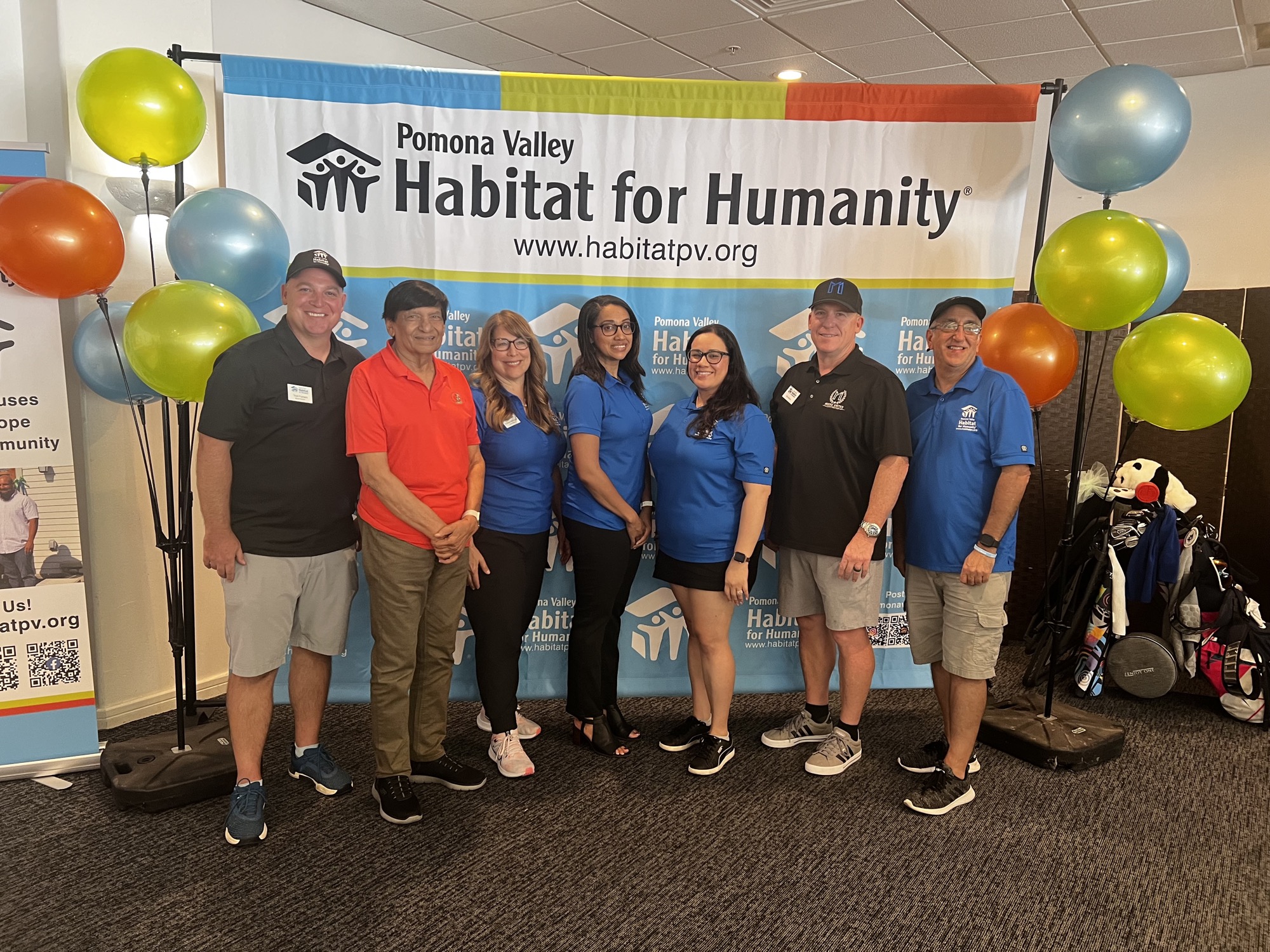 A group of people are gathered indoors, posing for a photo with colorful balloons. They are dressed in casual clothing and all have smiles on their faces, creating a cheerful atmosphere. This image is associated with Pomona Valley Habitat for Humanity, suggesting a connection to community activities or events.