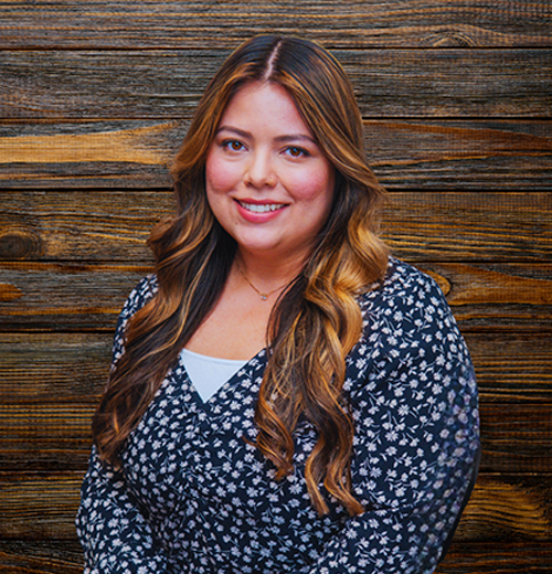 A woman with long, brown layered hair is smiling while posing against a wooden backdrop.