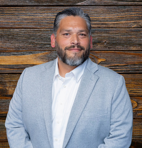 A man with a beard wearing a dress shirt and blazer, smiling. He is indoors with wooden elements in the background, captured in a headshot portrait.