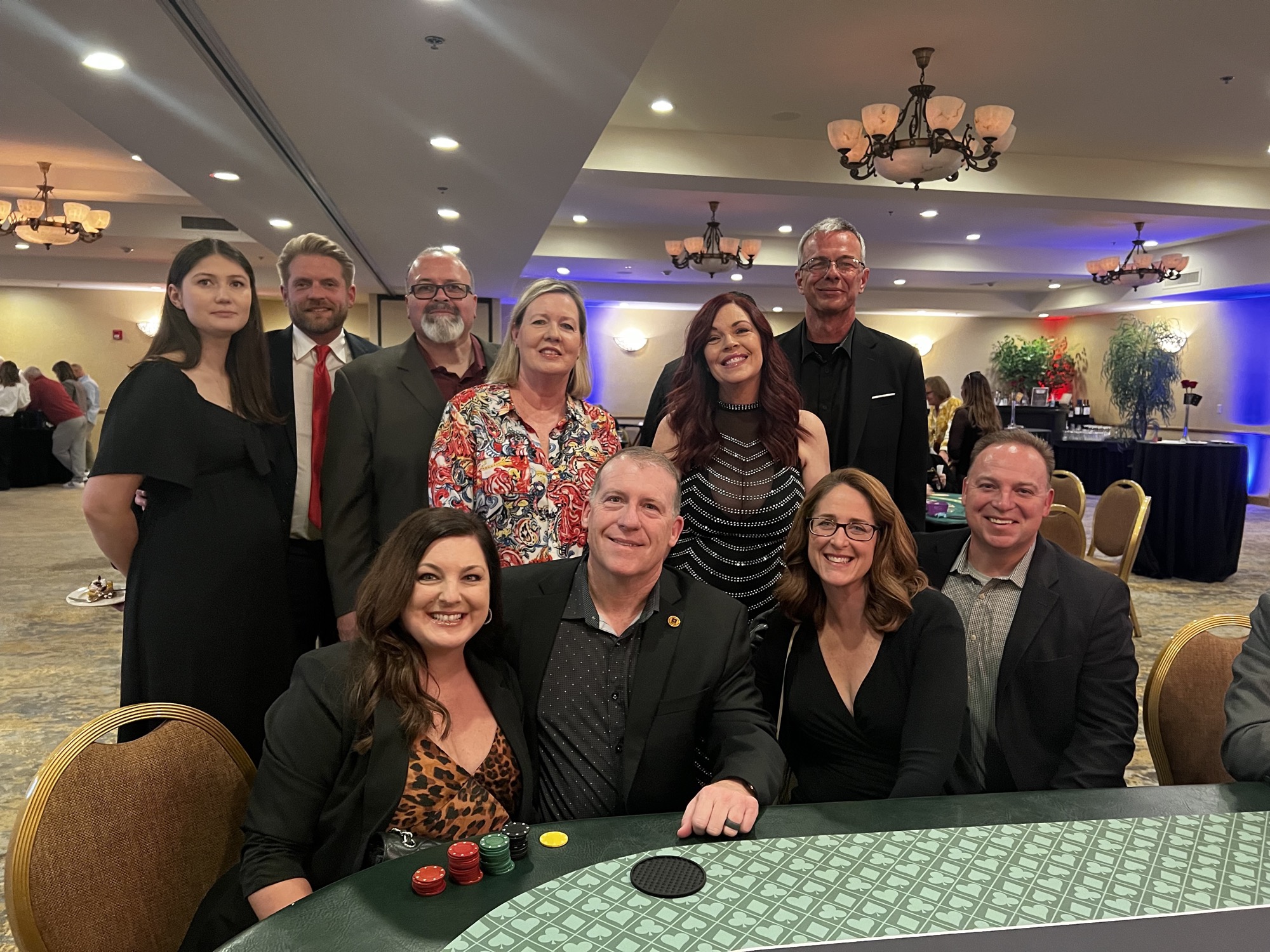 A group of people smiling and posing together for a photo indoors, with a table in front of them. The room has a ceiling and walls decorated, creating a party atmosphere.