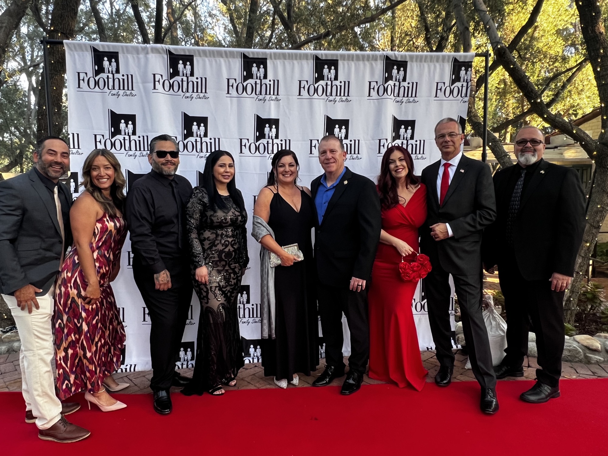 A group of people smiling and posing for a photo outdoors, standing on a red carpet with trees in the background. They are dressed in various outfits, including suits and dresses.