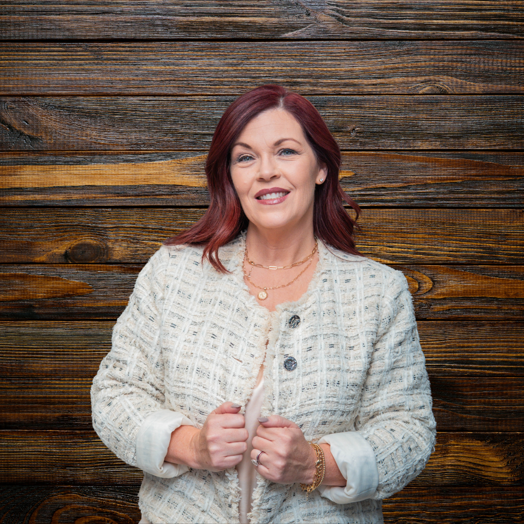 A woman smiling with her hands crossed, standing indoors in front of a wooden background.