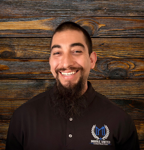 A man with a beard is smiling for the camera, wearing a shirt. The photo is taken indoors, capturing his face and upper body in a headshot style.