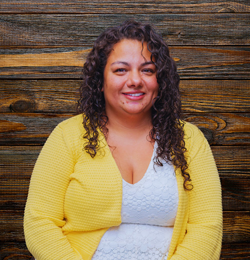 A person with curly hair is posing outdoors. The individual is a woman who is smiling and wearing clothing in yellow. The setting appears to include wooden elements, contributing to the overall ambiance of the photo.