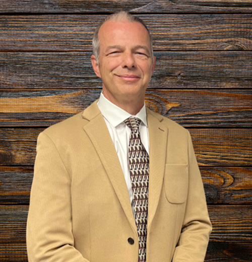 A man in a suit and tie stands indoors, smiling for the camera. He has a blazer over a dress shirt, with the collar visible. The setting features wooden elements in the background.