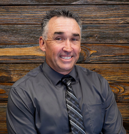 A man in a suit and tie smiles, showcasing his face, with a well-groomed appearance and a wooden backdrop.