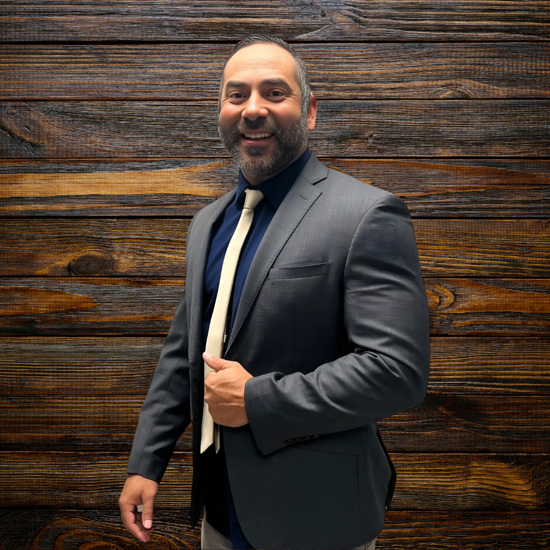 A man in a suit and tie, smiling while standing indoors. He has a beard and is wearing a blazer over a dress shirt. The background features wooden elements.