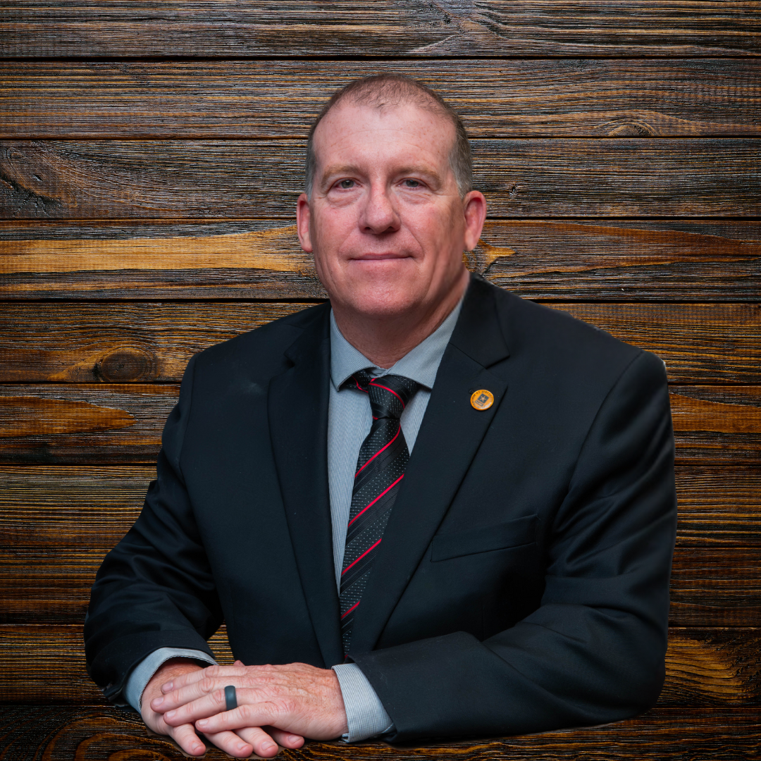 A man in a suit smiling at the camera, wearing a dress shirt and tie. He is indoors with a wooden background.